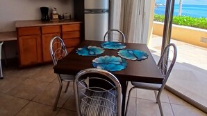 A dining area with a table set for four, surrounded by metal chairs, a kitchen area with wooden cabinets and a refrigerator, and a view of the ocean through a large sliding glass door.