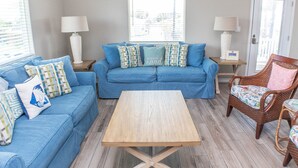 A living room with a blue sofa, a wooden coffee table, wicker chairs, and a view of the outside through a window. - JoJo's Heaven - in town Port Aransas, private pool. 4 BR/3bath. Perfect getaway! (Port Aransas)