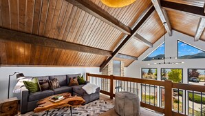 Una sala de estar moderna con techo de madera, un sofá seccional gris, un tapete con estampado de espiga y vistas al exterior a través de amplios ventanales.
