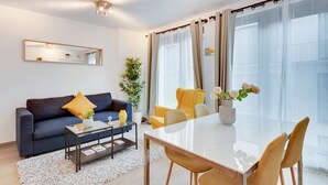 Een moderne woonkamer met een bank, salontafel, eettafel, stoelen en een groot raam met gordijnen.