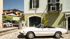 Une voiture décapotable blanche est garée devant un immeuble dont la porte est verte et qui possède un balcon orné de plantes en pot.