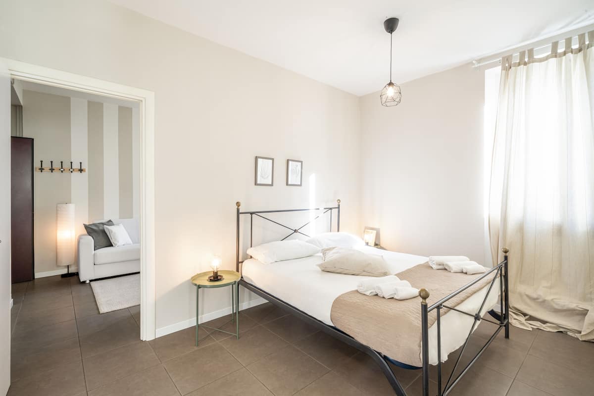 Makuuhuone, jossa on sänky, yöpöydät, lamppu, ikkuna verhoilla ja ovi, joka johtaa toiseen huoneeseen.