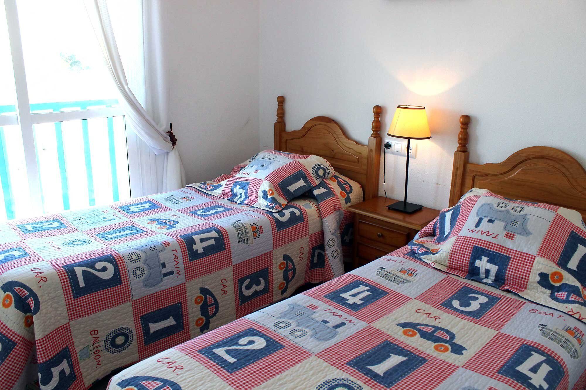 Une chambre avec deux lits, chacun recouvert d’une courtepointe ornée de motifs représentant des trains et des voitures. Une table de chevet avec une lampe se trouve entre les deux lits.
