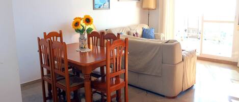 A dining area with a wooden table and chairs, a vase with sunflowers, a sofa with cushions, and a lamp.