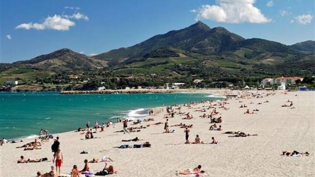 A beach with people sunbathing and swimming, a mountain in the background, and a clear sky.