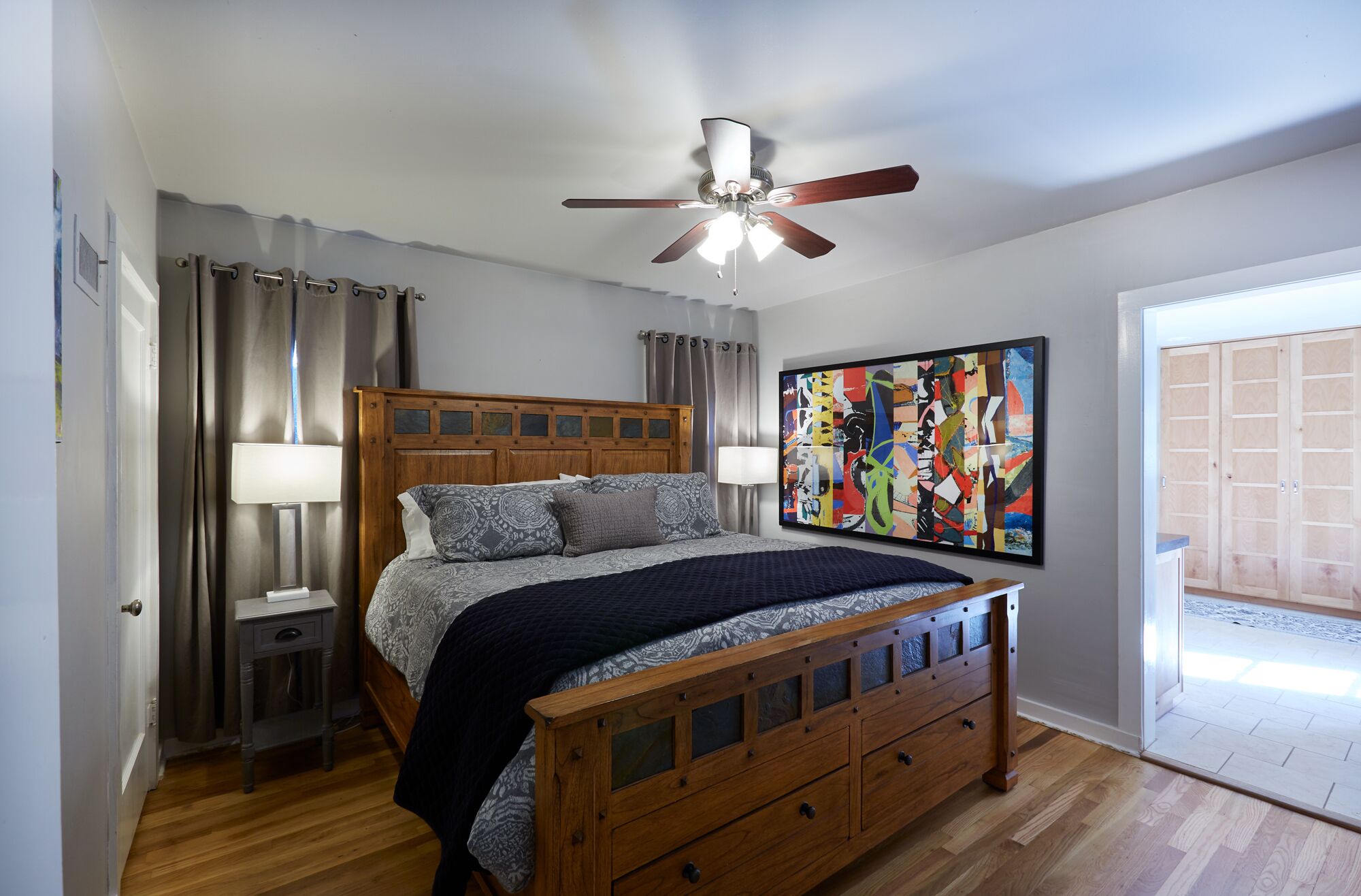 A bedroom with a wooden bed, a ceiling fan, a large abstract painting, and a view of the outside.