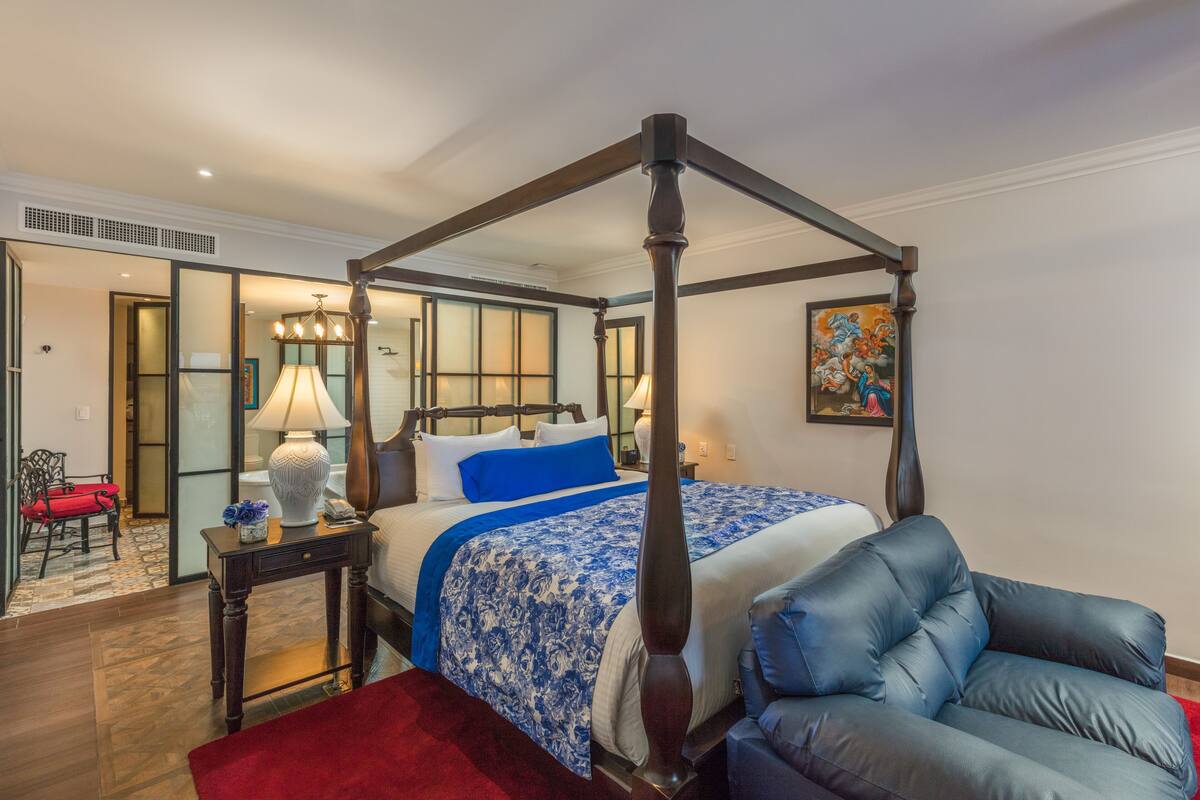 A four-poster bed with a blue and white bedspread, a red throw blanket, and a blue upholstered chair.