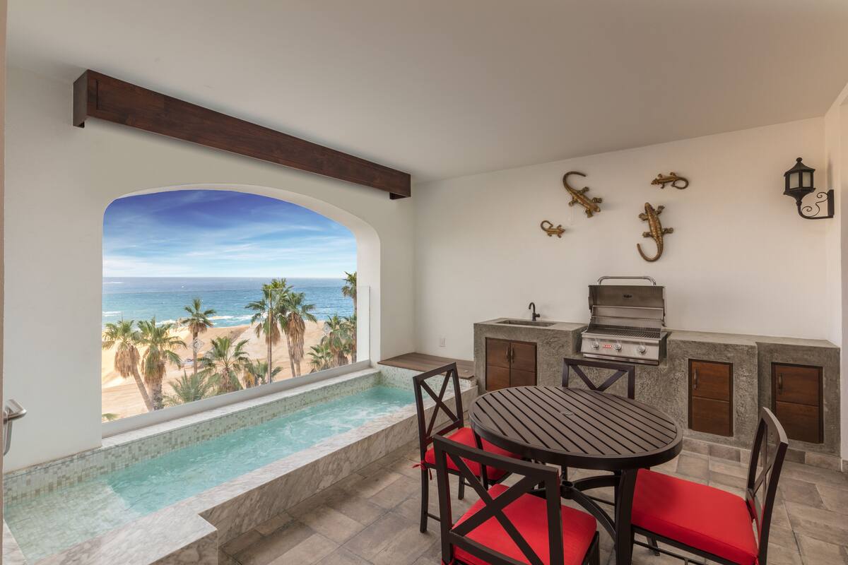 A balcony with a view of the beach and ocean, a pool, a grill, and a table with red chairs.