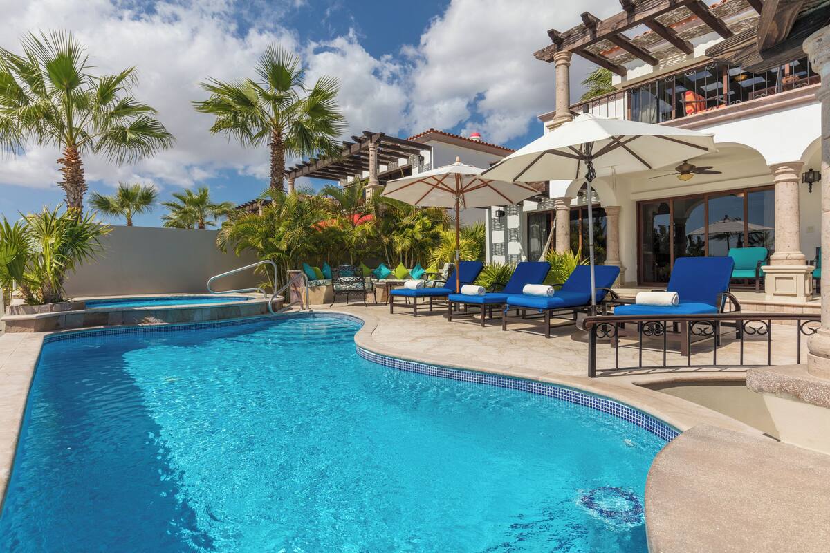 A swimming pool with blue water, surrounded by lounge chairs and umbrellas, palm trees, and a building with a balcony.