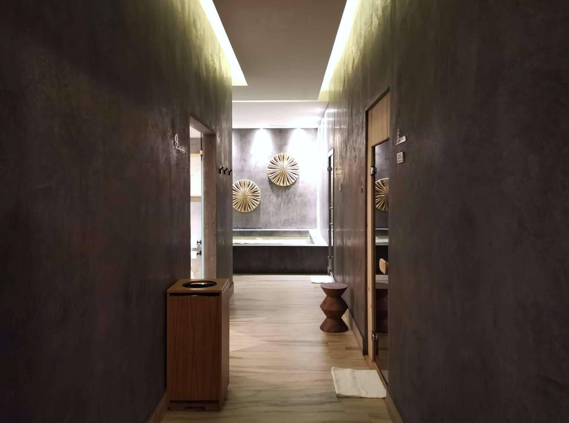 A hallway with a trash bin, a wooden stool, and a wall-mounted toilet.