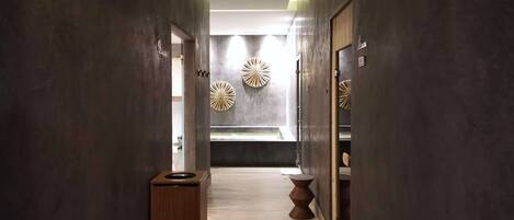 A hallway with a trash bin, a wooden stool, and a wall-mounted toilet.