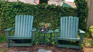 Two green Adirondack chairs with a small table and potted plants between them, set on a stone pathway.