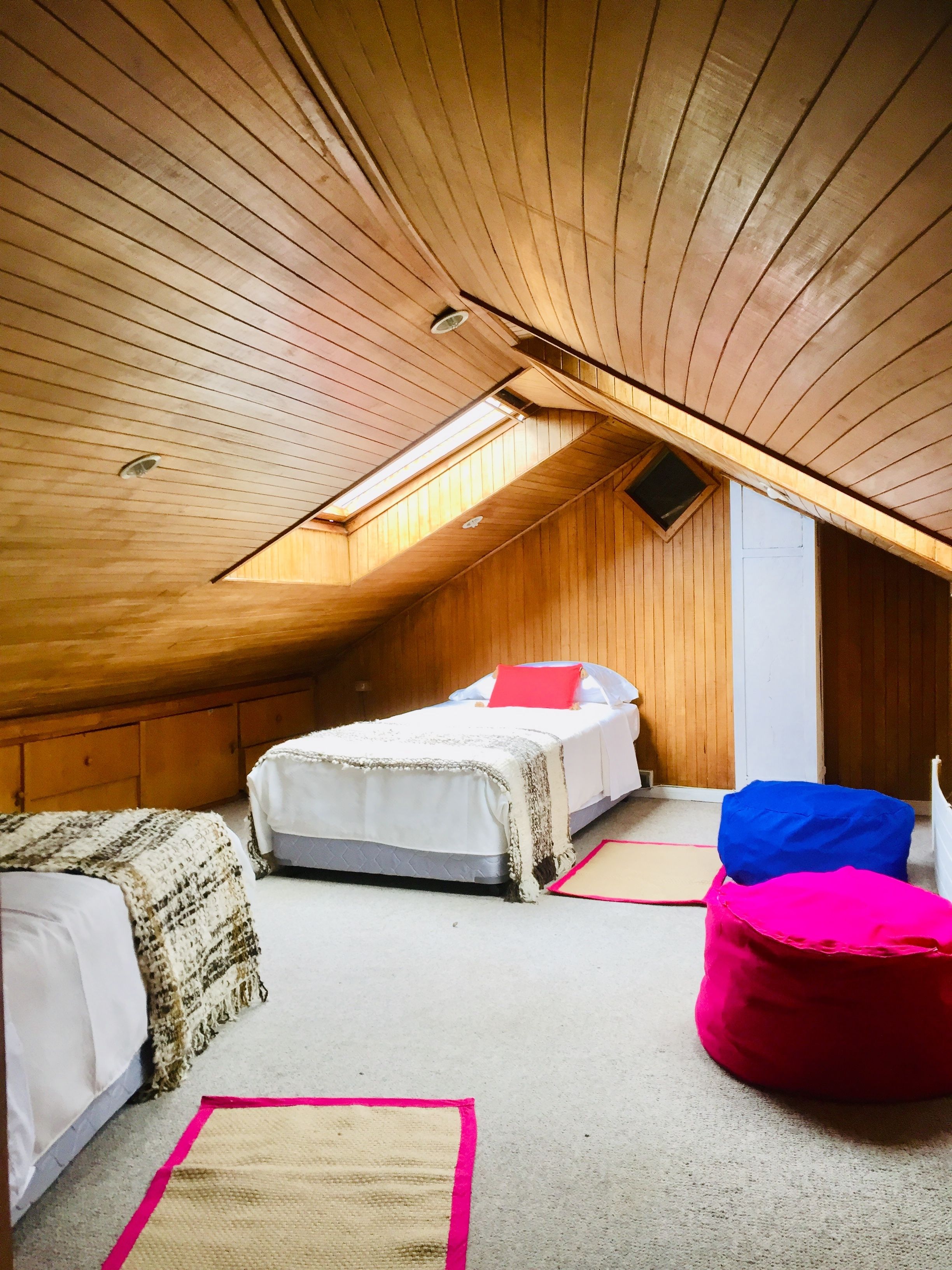Un dormitorio abrigado en el ático, con techo inclinado, dos camas, un sillón de peluche rojo y un tragaluz pequeño.