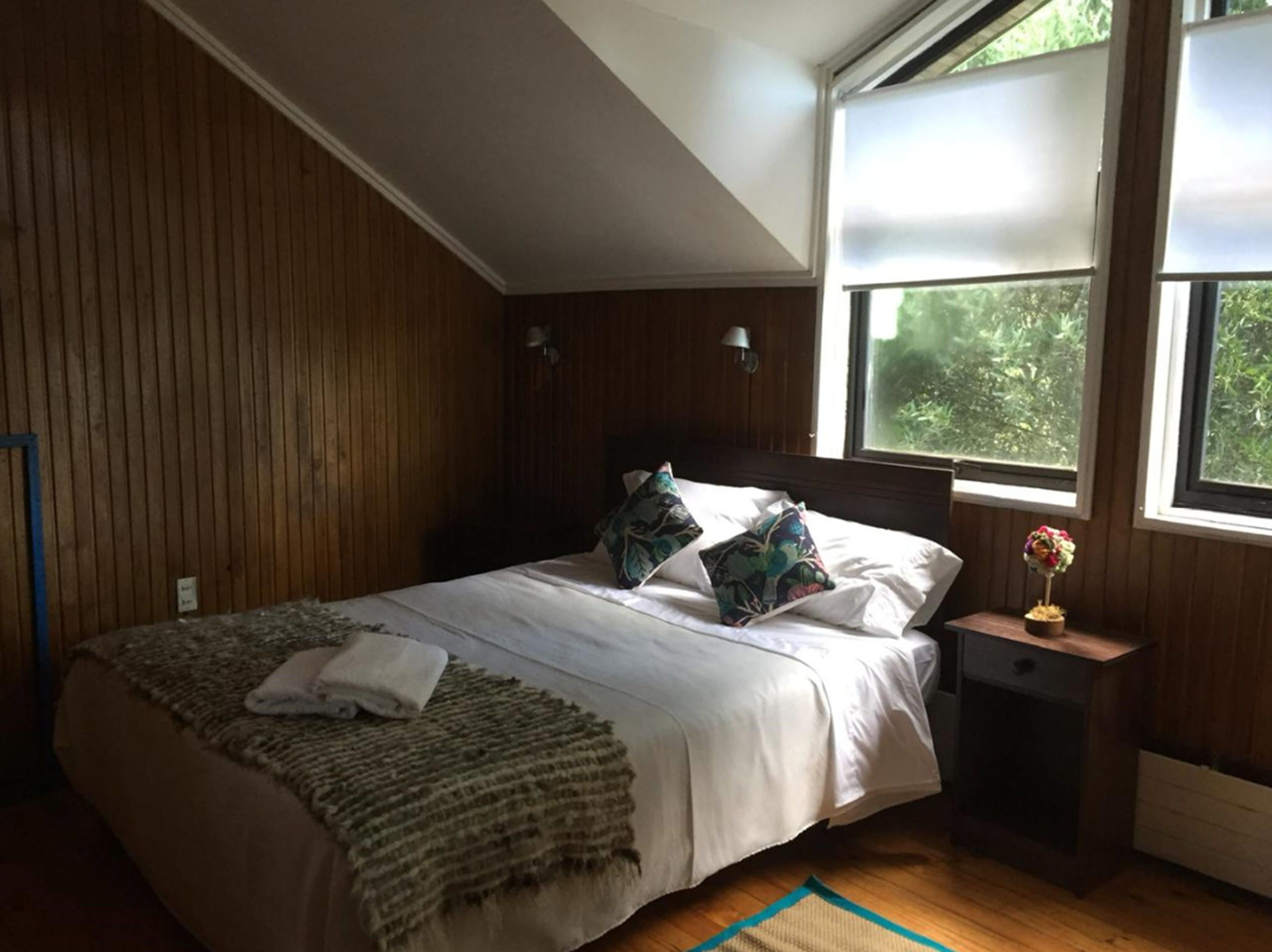 Un dormitorio con pared de paneles de madera, una cama con sábanas blancas, dos almohadas, una mesita de noche con un florero y una ventana con vistas a la vegetación.