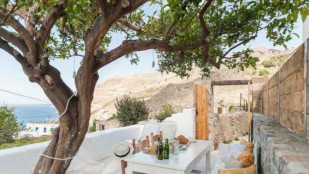 A terrace with a white table, chairs, and a view of the sea and hills.