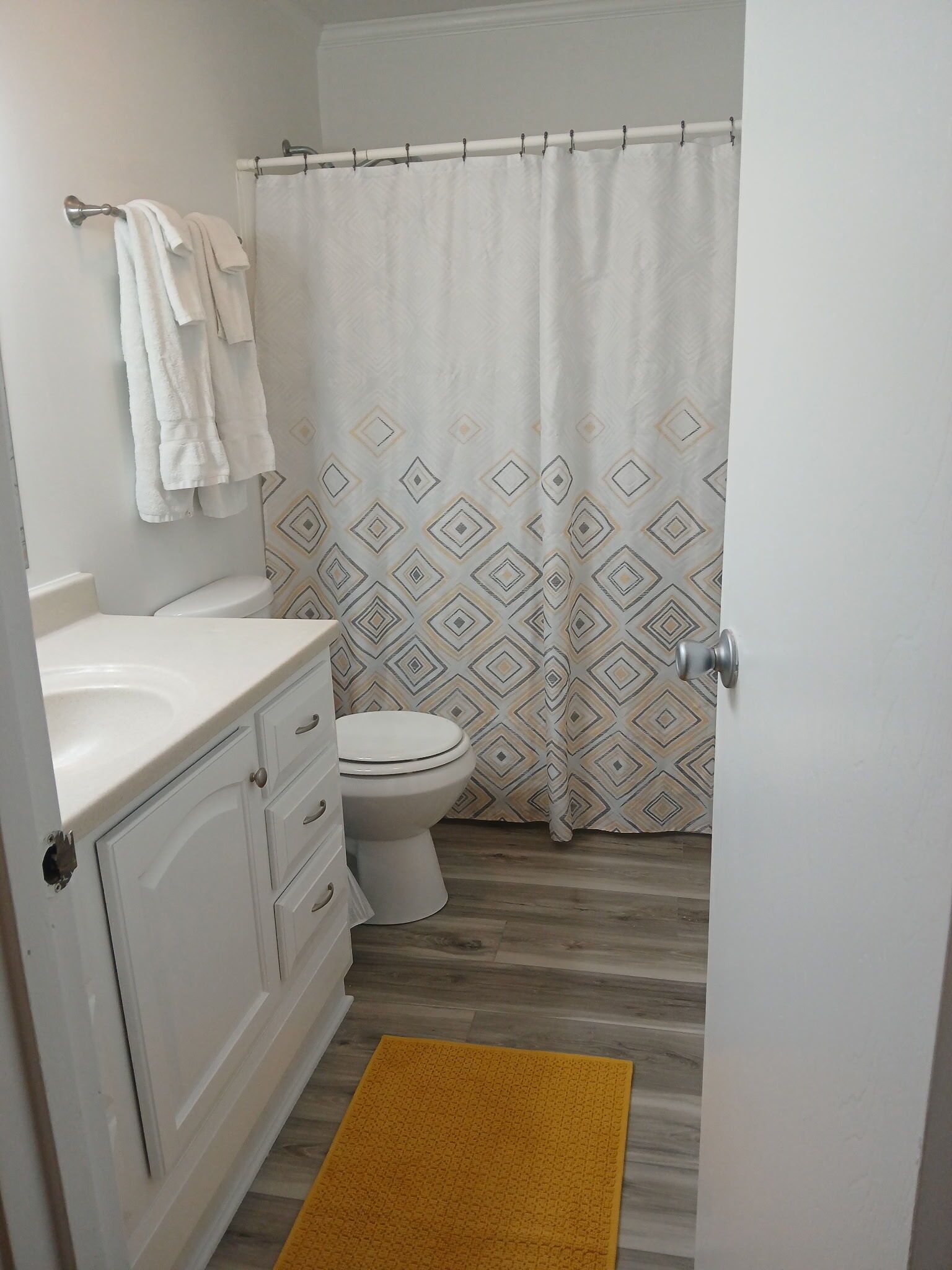 Un baño con un inodoro blanco, un lavamanos con encimera blanca y una cortina de ducha de estampado geométrico. Hay una alfombra amarilla en el suelo.
