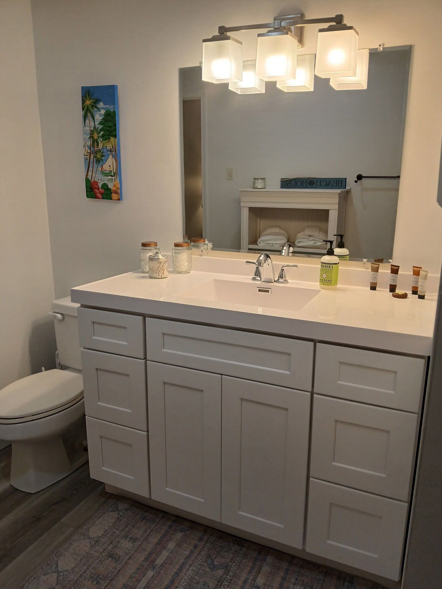 A bathroom with a white vanity, a large mirror, and a toilet. There are jars on the vanity and a bottle of soap on the counter.