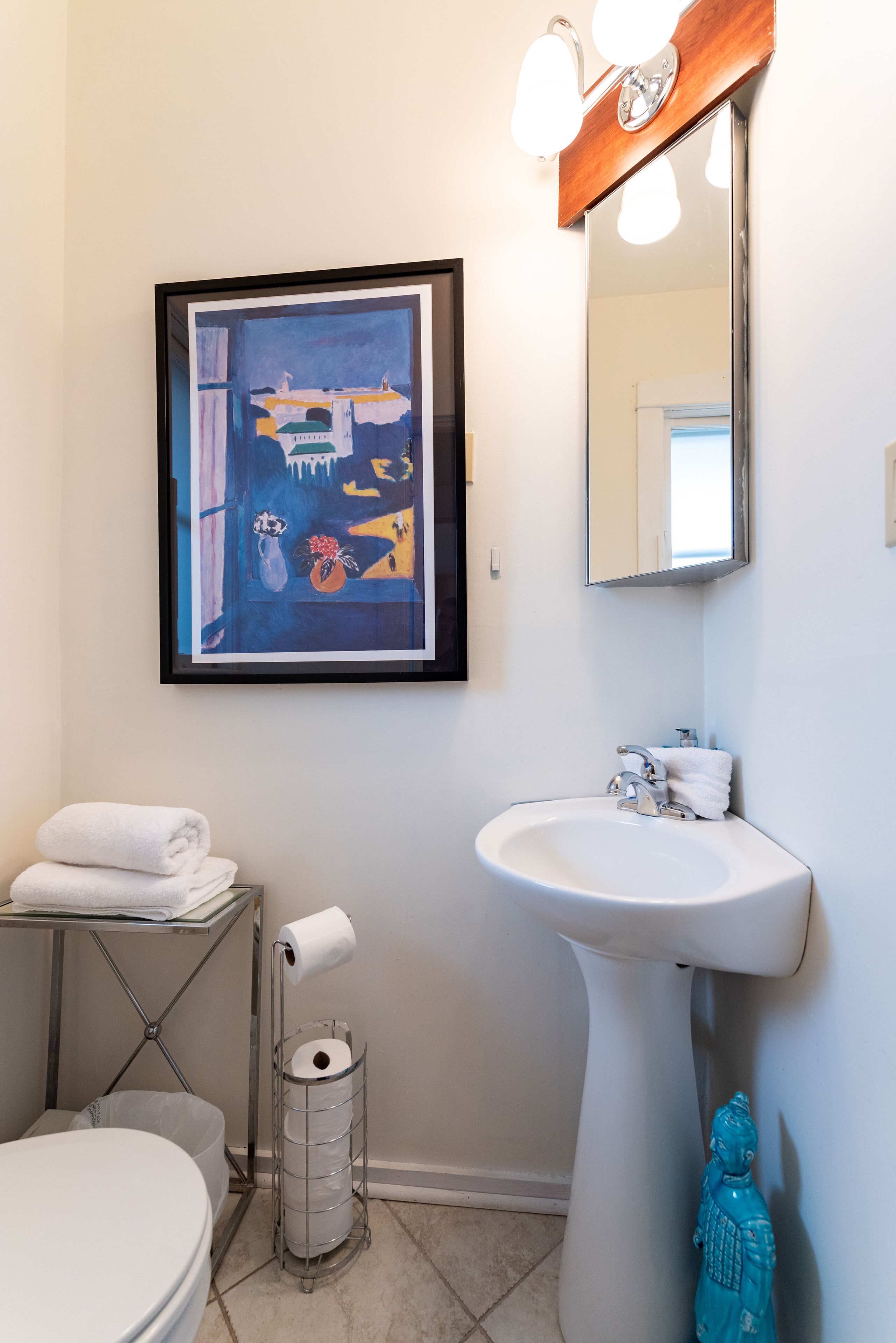 Et badeværelse med et indrammet abstrakt billede, en vask, et toilet og en blå dekorativ figur.