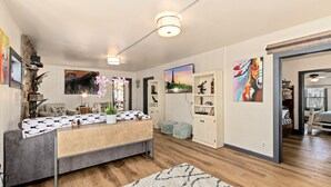 Una sala de estar moderna con un sofá gris, una mesa de centro de madera y un tapete blanco. Se observan dos lámparas colgantes, una estantería y un cuadro de un paisaje urbano.