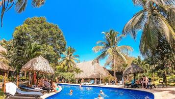 Piscina rodeada de palmeras y cabañas de techo de paja, con sillones de descanso y gente nadando.