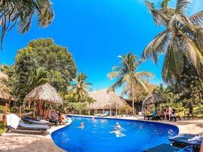 A swimming pool surrounded by palm trees and thatched-roof huts, with lounge chairs and people swimming. - Cabañas Palomino I (Dibulla)