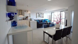 A modern kitchen with white cabinetry, a dining area with a white table and black chairs, a blue sofa, and a large window. - Blue Coral Terrace, Coast Apartment 31 (The Entrance)