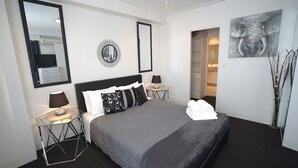 A hotel room with a bed, bedside tables, a mirror, a wall art piece, and a door leading to a bathroom. - Blue Coral Terrace, Coast Apartment 31 (The Entrance)