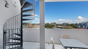 A balcony with a spiral staircase, a table, and chairs, overlooking a cityscape with buildings and a clear sky. - Blue Coral Terrace, Coast Apartment 31 (The Entrance)