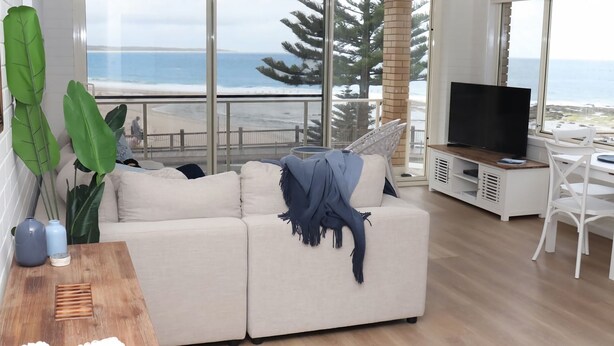 Una sala de estar moderna con un gran ventanal con vistas al océano, un sofá blanco, un mueble para televisión y un comedor.