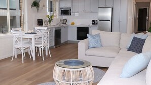 Una cocina moderna con armarios blancos, un comedor con sillas blancas y una sala de estar con un sofá color beige y cojines estampados.