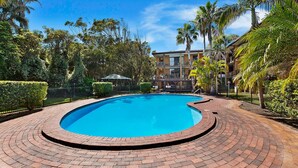Piscina com borda de tijolos, cercada por vegetação e um edifício com varandas.