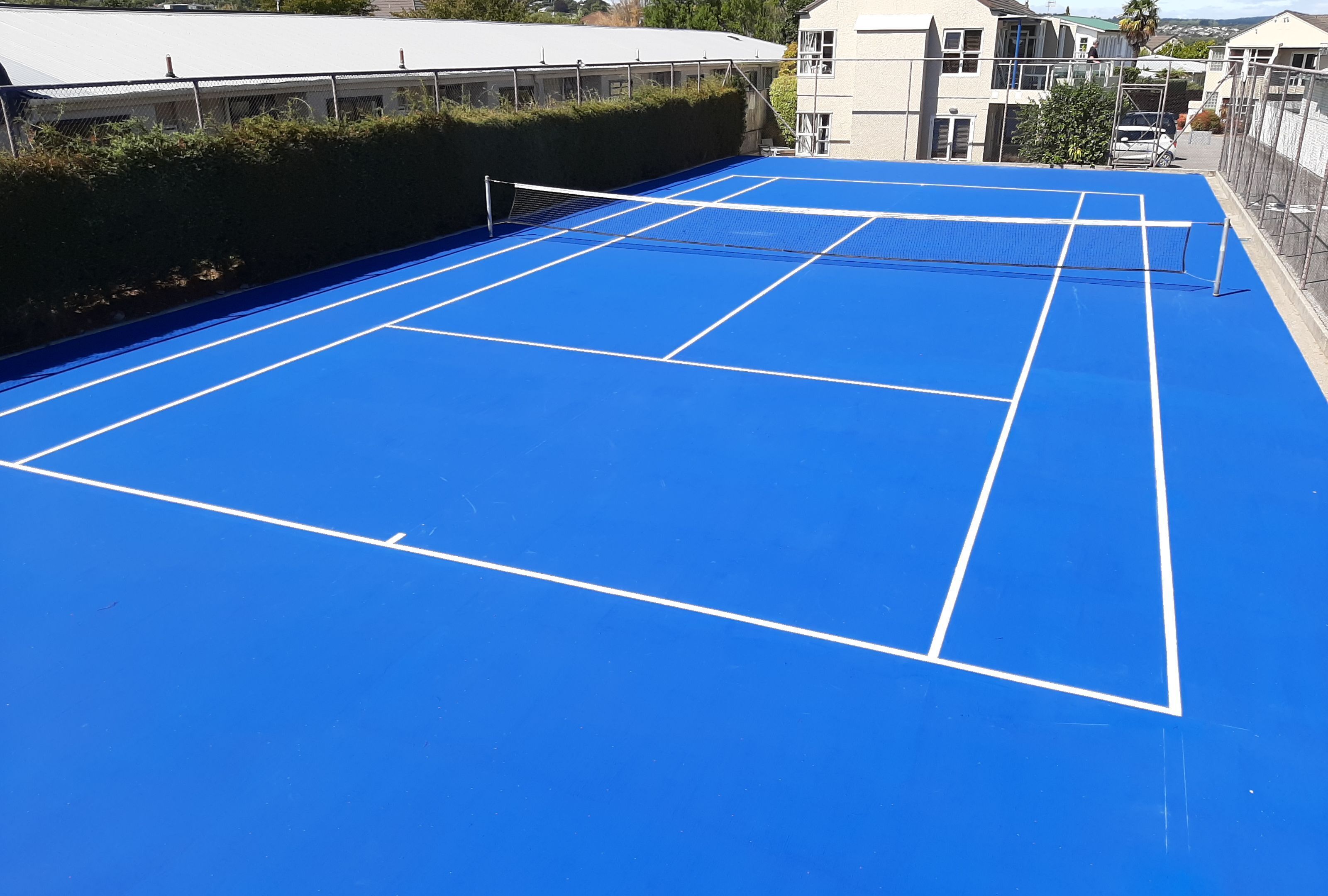 A blue tennis court with white boundary lines, surrounded by a fence and buildings in the background.