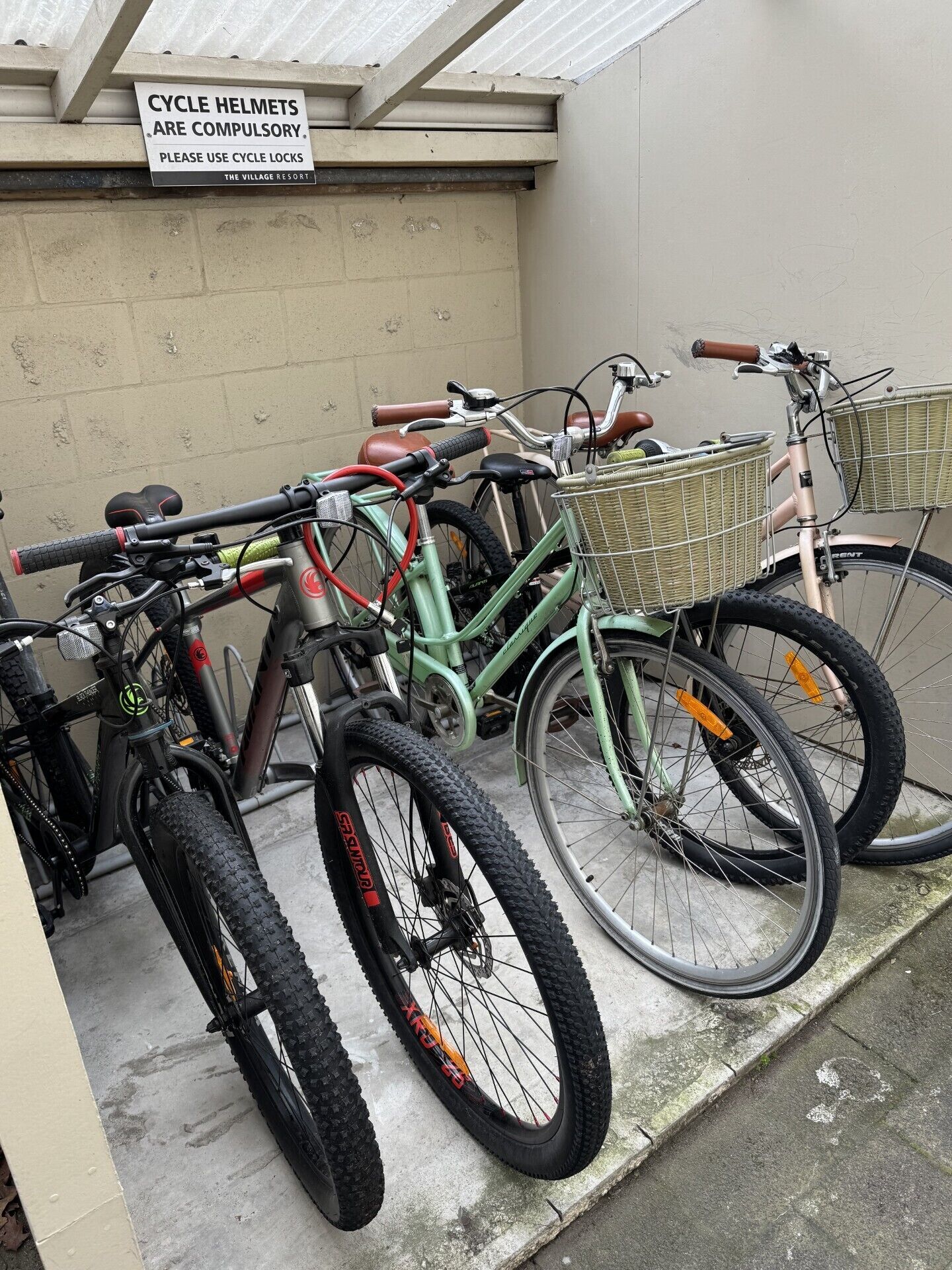 A bicycle parking area with several bicycles, including mountain bikes and a green road bike, with baskets attached. A sign above indicates that cycle helmets are compulsory and cycle locks should be used.