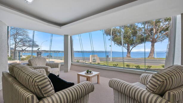 A modern living room with striped sofas and a small table, large windows with a view of the sea and trees. - The Village Resort (Taupo)