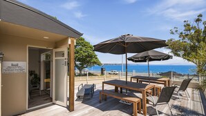 A wooden deck with tables and chairs, a large umbrella, and a view of the sea. - The Village Resort (Taupo)