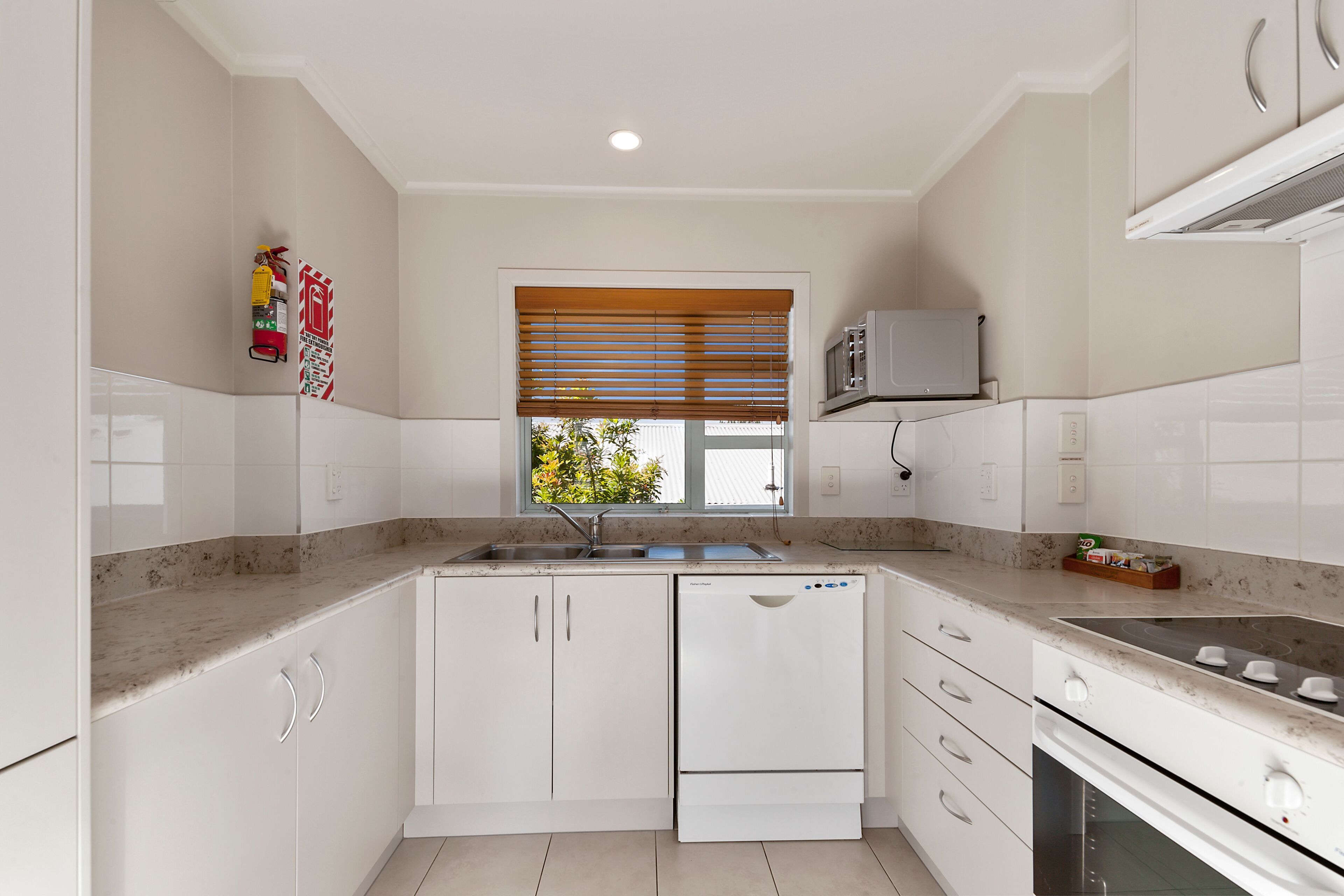 A modern kitchen with white cabinetry, a built-in oven, a microwave, and a window with blinds.