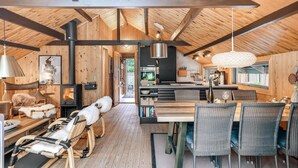 Interior acolhedor de uma cabana com teto de madeira, lareira, área de jantar com cadeiras de vime e cozinha com eletrodomésticos modernos.