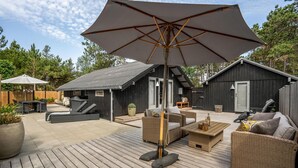 Um deck de madeira com móveis de exterior, um grande guarda-sol e um edifício pequeno com fachada escura.