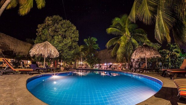Un área de piscina iluminada de noche con sillones de descanso, palmeras y sombrillas de paja.