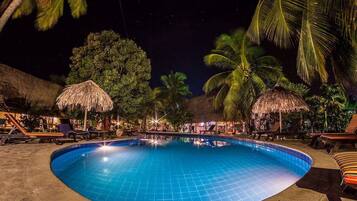 Un área de piscina iluminada de noche con sillones de descanso, palmeras y sombrillas de paja.