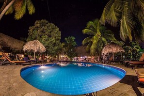 Un área de piscina iluminada de noche con sillones de descanso, palmeras y sombrillas de paja.