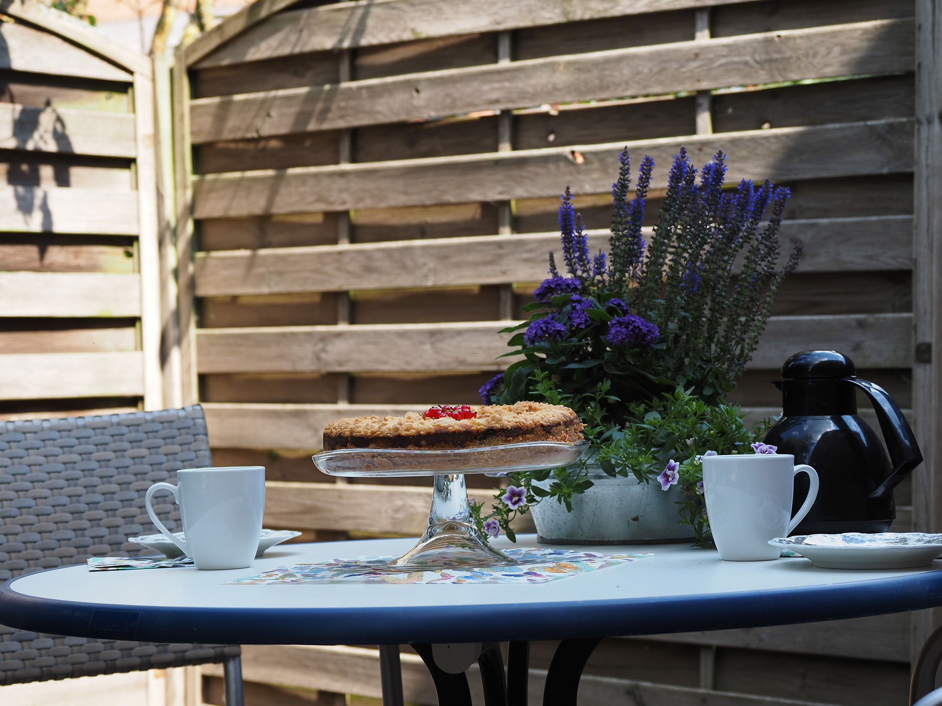 A round table with a glass cake stand, two white cups, a coffee pot, and a floral tablecloth.