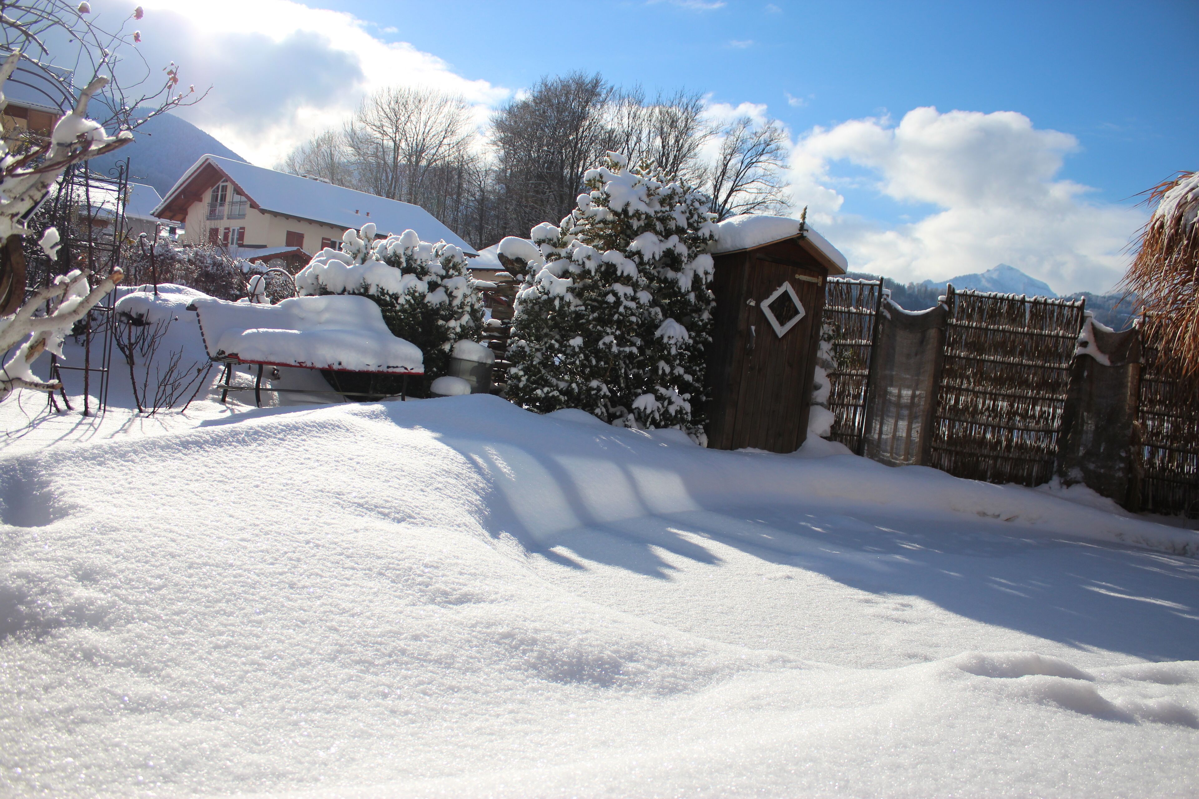 A snow-covered landscape with a wooden fence, a small wooden structure, and a house with a snow-covered roof.