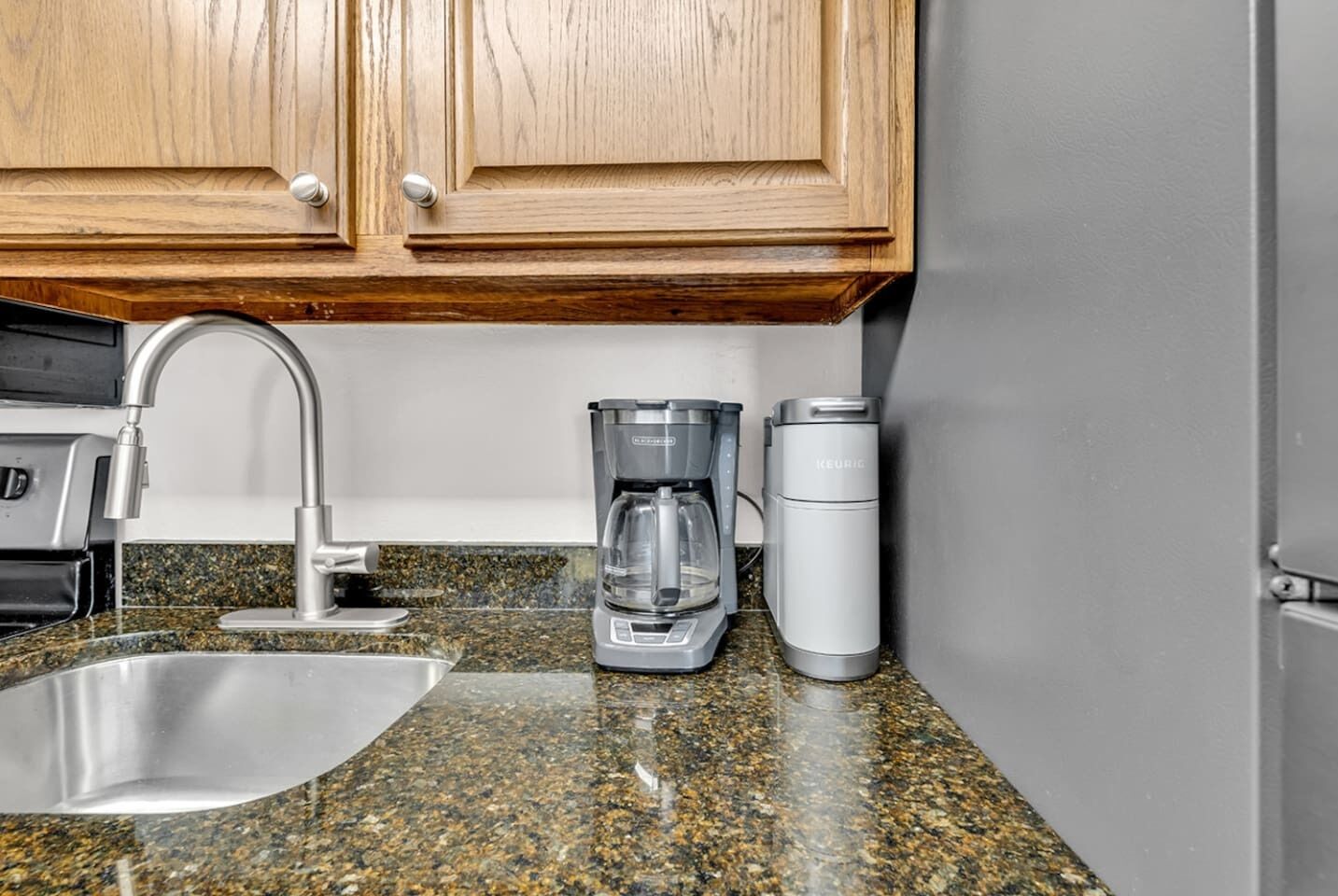 A kitchen with a granite countertop, stainless steel sink, wooden cabinets, a coffee maker, and a Keurig coffee pod dispenser.