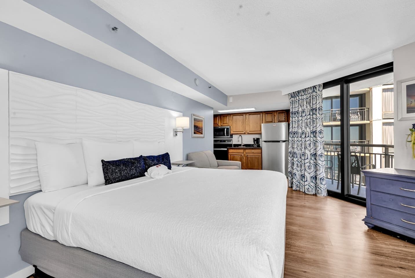 A modern hotel room with a large bed, a kitchenette, and a balcony with patterned curtains.