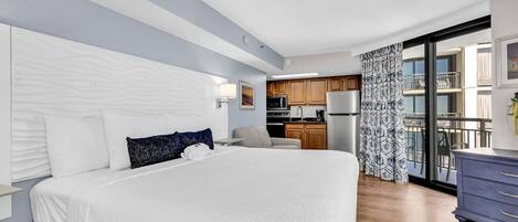 A modern hotel room with a large bed, a kitchenette, and a balcony with patterned curtains.