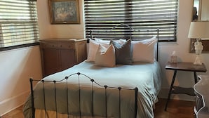 Um quarto com estrutura de cama de metal, piso de madeira, cômoda, mesa de cabeceira e um quadro na parede.