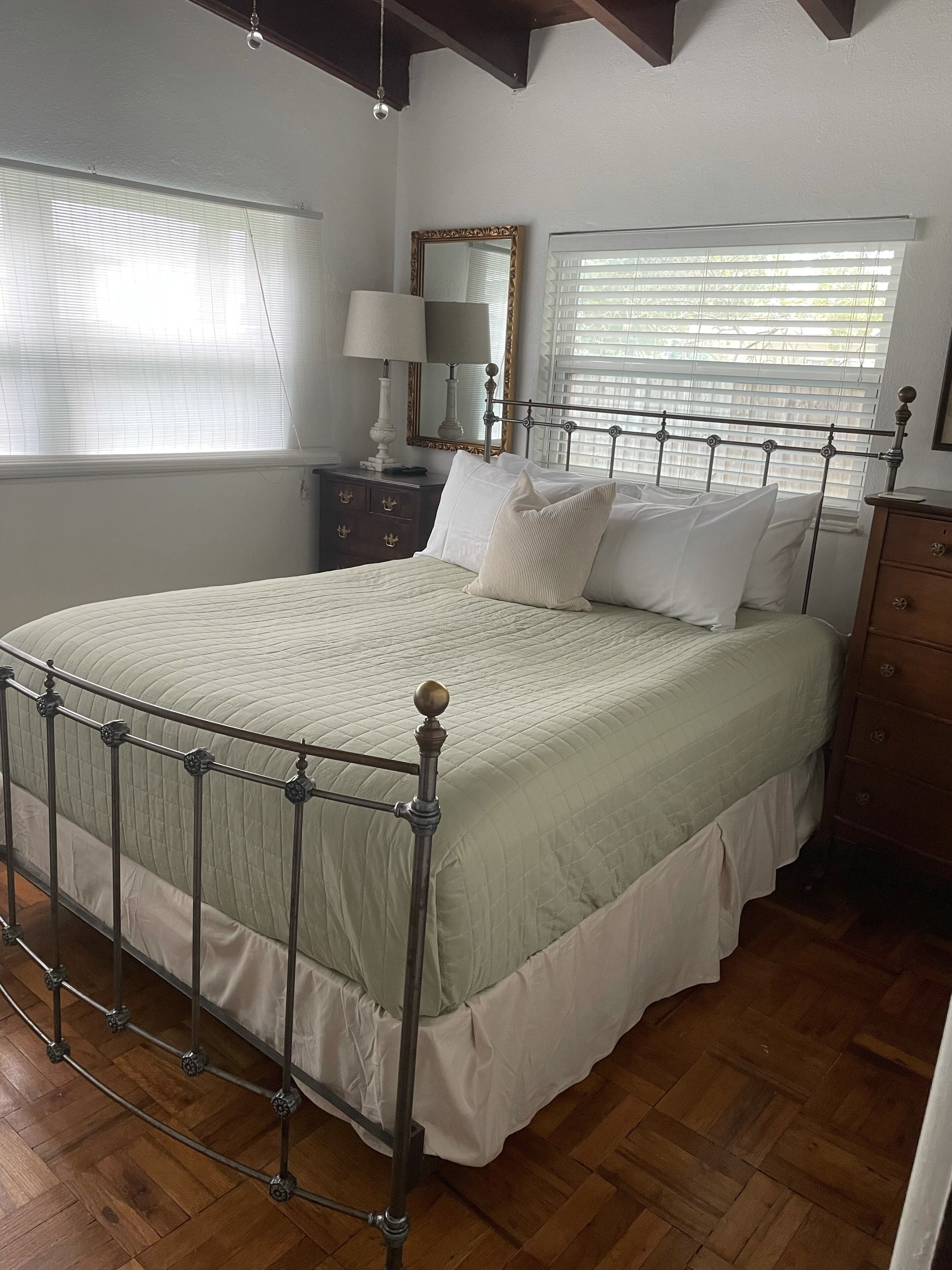 Une chambre à coucher bien rangée, avec un lit à ossature métallique, une commode en bois, une table de chevet avec une lampe et un grand miroir au mur.