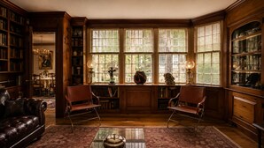 A library with bookshelves, two leather chairs, a glass coffee table, and a bay window with blinds. - 29 India House (Nantucket)