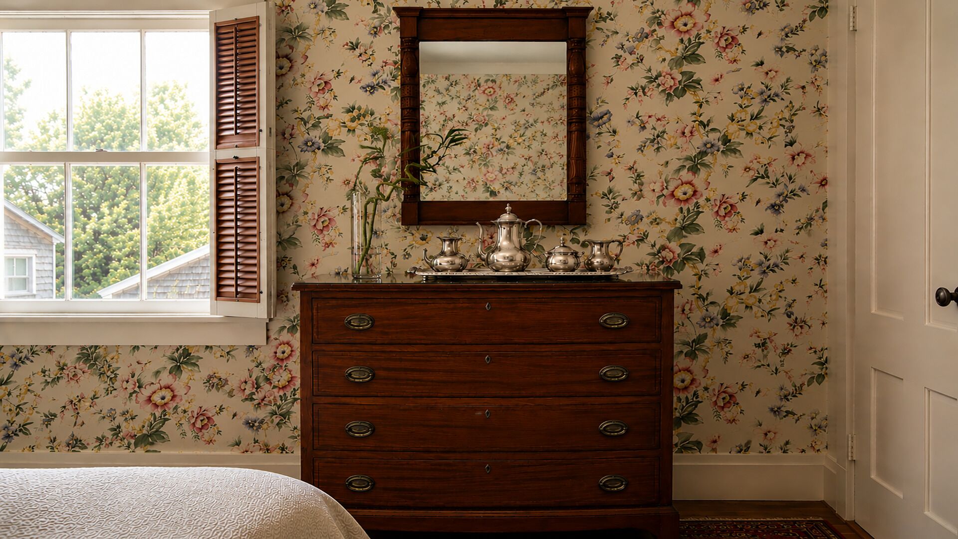 A room with a wooden dresser, floral wallpaper, a mirror, and a window with shutters.